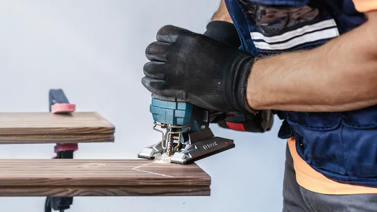 Person wearing safety equipment cuts wood with a jigsaw.