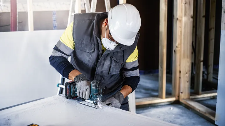 A person wearing safety equipment cuts drywall with a jigsaw.