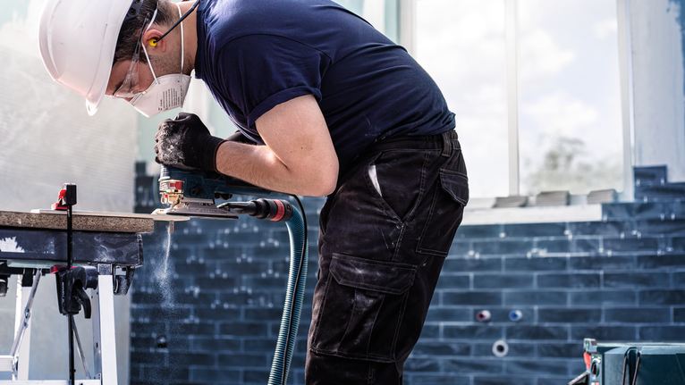 A person wearing safety equipment uses a power tool to cut a tile.