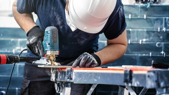 Person wearing safety equipment uses a jigsaw to cut a tile on a workbench.