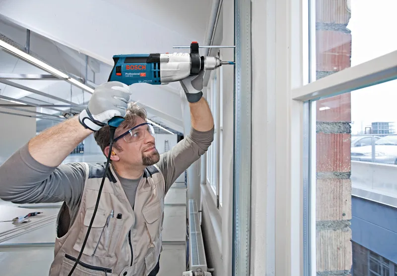 A person wearing safety equipment drills into a wall frame near a window.