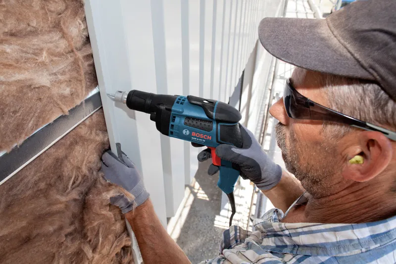 A person wearing safety equipment uses a drywall screwdriver to fasten metal siding.