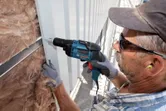 A person wearing safety equipment uses a drywall screwdriver to fasten metal siding.