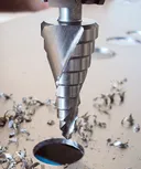 Burghiu în trepte care creează o gaură într-o tablă metalică, înconjurat de așchii metalice.