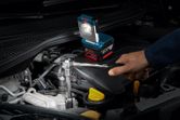 O persoană lucrează la motorul unei mașini folosind o cheie franceză, având în apropiere o lanternă de lucru fără fir.