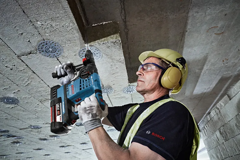 O persoană purtând echipament de siguranță găurește un tavan de beton cu un ciocan rotopercutor fără fir.