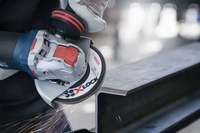 O persoană care poartă echipament de siguranță șlefuiește o grindă metalică cu o unealtă electrică, creând scântei.