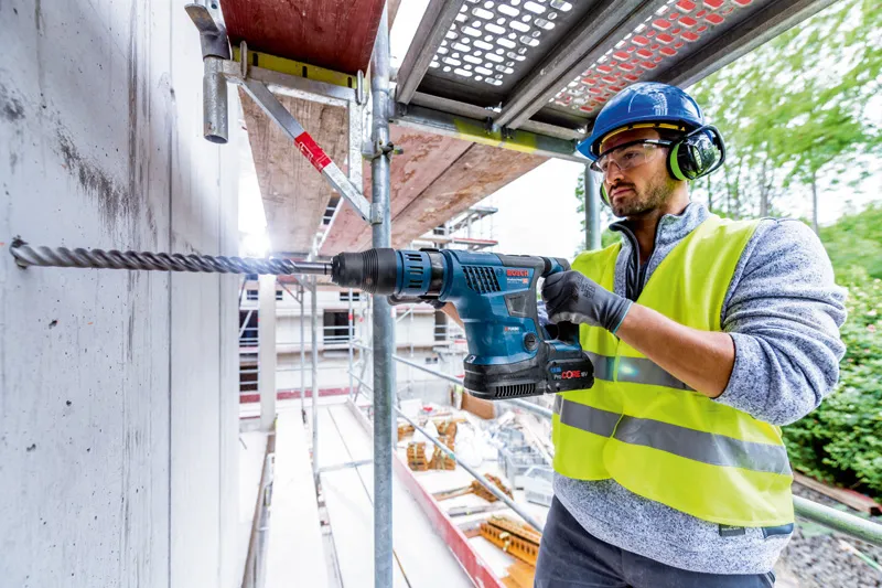 O persoană purtând echipament de siguranță găurește în beton folosind un ciocan rotopercutor fără fir.