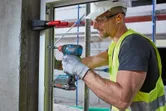 O persoană purtând echipament de siguranță fixează rama unei ferestre cu o bormașină fără fir.