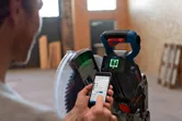 O persoană ajustează un ferăstrău circular fără fir cu un smartphone într-un atelier interior.