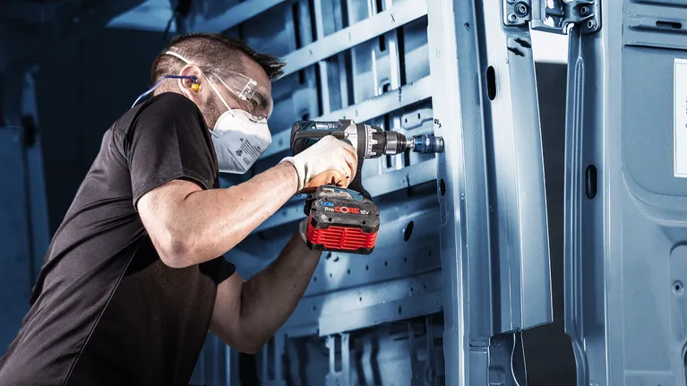 O persoană care poartă echipament de siguranță folosește o bormașină fără fir pentru a lucra la o structură metalică.