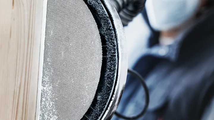 Persoană purtând echipament de siguranță șlefuind o suprafață de lemn cu o unealtă electrică.