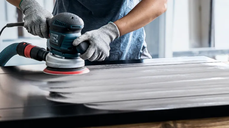 Persoană care poartă echipament de siguranță șlefuiește o suprafață de lemn cu o unealtă electrică.