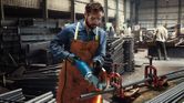 O persoană purtând echipament de siguranță taie o țeavă metalică cu o unealtă electrică într-un atelier industrial.