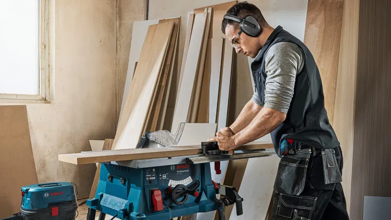 O persoană purtând echipament de siguranță taie lemne cu un ferăstrău circular într-un atelier.