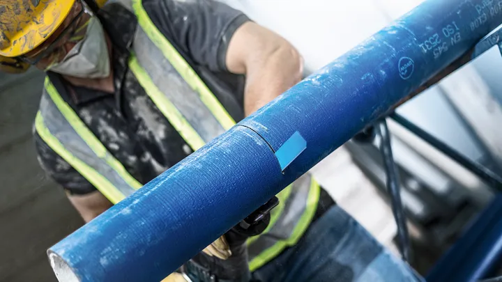 O persoană care poartă echipament de siguranță taie o țeavă albastră cu o unealtă electrică.