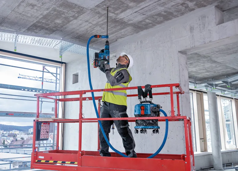 O persoană purtând echipament de siguranță găurește tavanul de beton al unui lift.