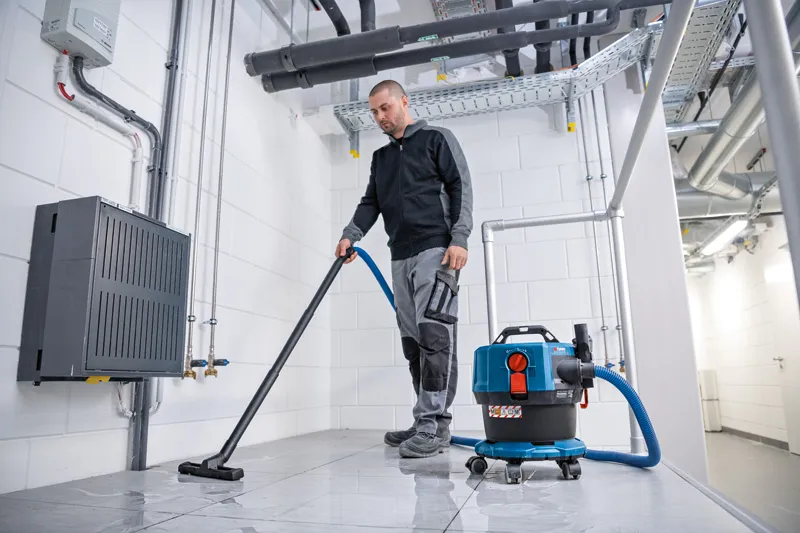 O persoană aspiră o podea cu gresie cu un aspirator umed și uscat într-o cameră industrială.