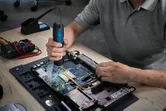 Persoană care folosește o șurubelniță fără fir pentru a asambla un laptop pe o bancă de lucru.