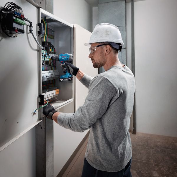 O persoană care poartă echipament de siguranță folosește o bormașină fără fir pe un panou electric.