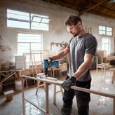 O persoană purtând echipament de siguranță găurește într-un cadru de lemn într-un atelier.