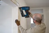 O persoană care poartă echipament de siguranță folosește o ciocan de cuie fără fir pe tocul unei uși.