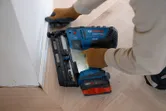 O persoană care poartă echipament de siguranță folosește o ciocan de cuie fără fir pentru a fixa marginea plintei.