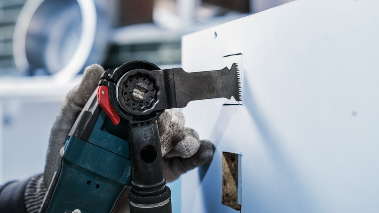 O persoană care poartă echipament de siguranță taie o gaură dreptunghiulară în gips-carton cu o unealtă electrică.