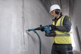 O persoană purtând echipament de siguranță găurește un perete de beton cu o unealtă electrică.