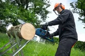 O persoană care poartă echipament de siguranță folosește un ferăstrău cu lanț fără fir pentru a tăia un buștean în aer liber.