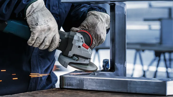 O persoană care poartă echipament de siguranță șlefuiește o grindă metalică cu un polizor unghiular.