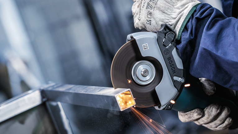 Persoană purtând echipament de siguranță tăind o grindă metalică cu o polizor unghiular.