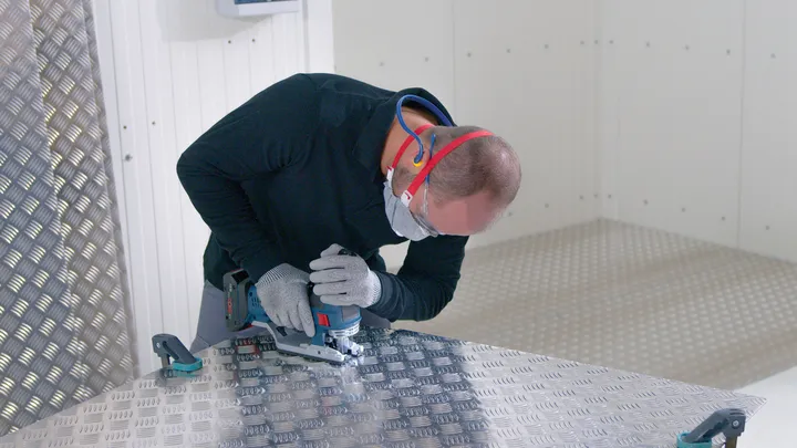 O persoană care poartă echipament de protecție folosește o unealtă electrică pentru a tăia o tablă de metal.