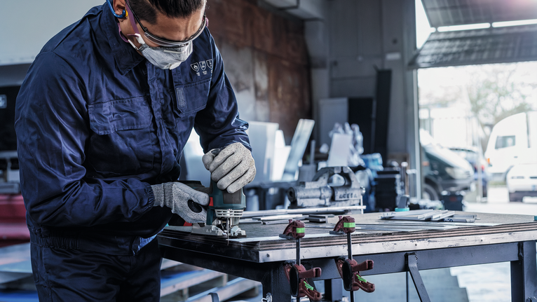 Persoană care poartă echipament de siguranță folosește o unealtă electrică pentru a șlefui metalul pe o bancă de lucru.