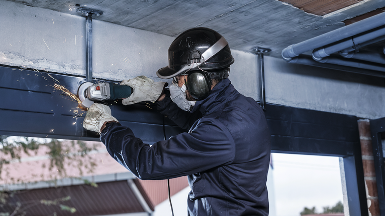 O persoană purtând echipament de siguranță șlefuiește o grindă metalică deasupra capului cu o unealtă electrică.