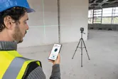 O persoană care poartă echipament de siguranță folosește un smartphone pentru a controla un instrument de nivelare cu laser pe un trepied.