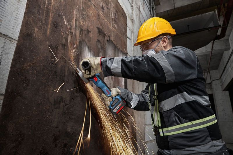 O persoană care poartă echipament de siguranță șlefuiește o tablă metalică ruginită cu un polizor unghiular fără fir.