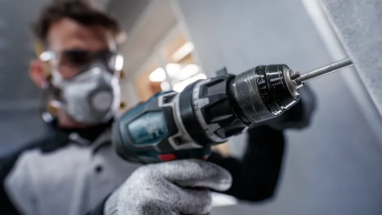 Persoană purtând echipament de siguranță găurește un perete cu o unealtă electrică.