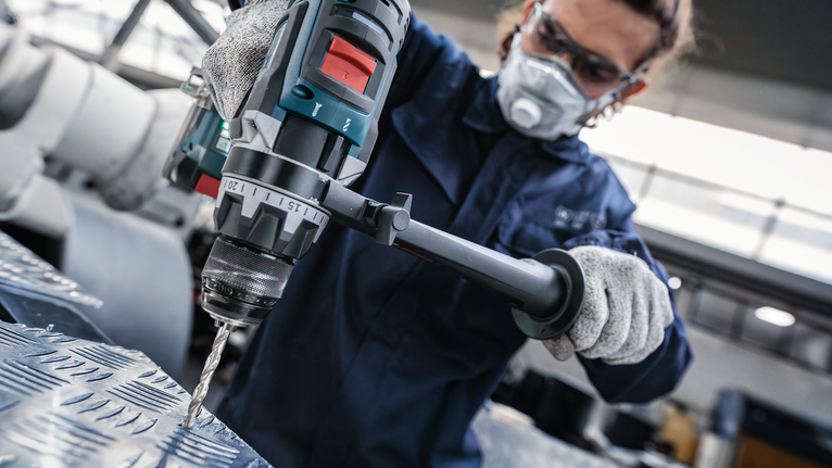 Persoană purtând echipament de siguranță găurește o tablă de metal într-un atelier industrial.