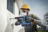 O persoană purtând echipament de siguranță găurește un perete exterior cu o unealtă electrică.