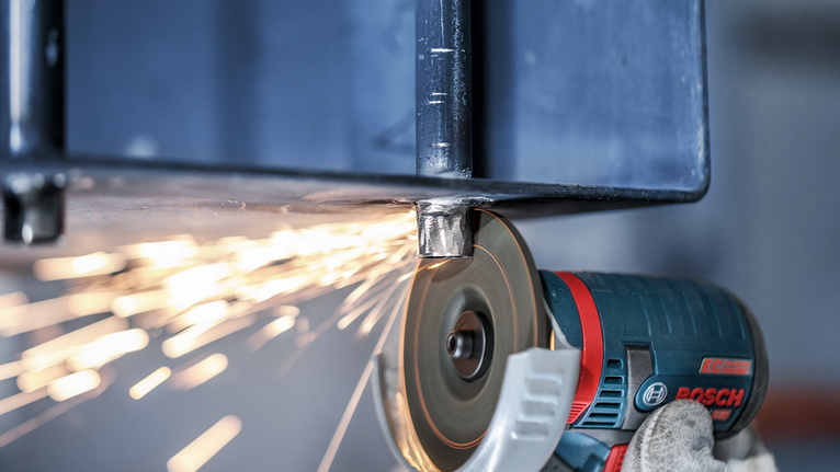 Un muncitor cu mănuși, purtând echipament de protecție, șlefuiește o grindă metalică cu un polizor unghiular, producând scântei.