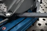 Fierăstrău de tăiat metal cu linie de ghidare roșie care taie o bară de oțel pe o bancă de lucru perforată.
