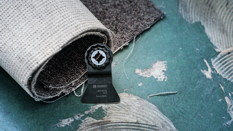 Lamă oscilantă a sculei pe podeaua verde lângă covorul rulat.