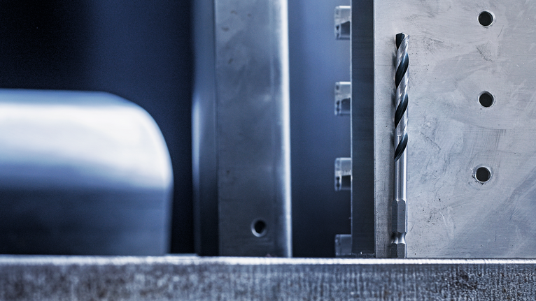 Burghiu metalic aliniat vertical lângă o suprafață metalică prelucrată cu găuri.