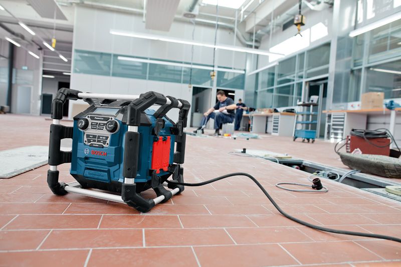 Radio de șantier pe o podea cu gresie într-un atelier, cu oameni care lucrează în fundal.