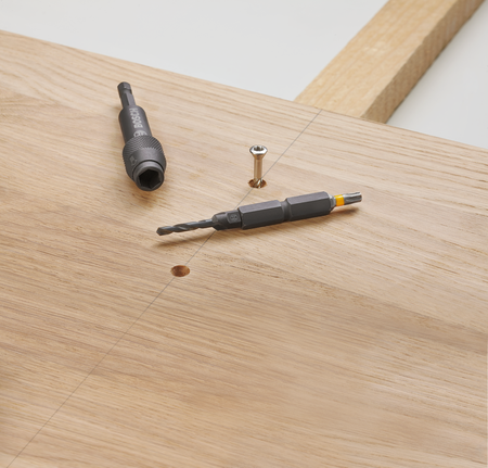 Бургија и одвијач леже на дрвеној дасци поред шрафа.