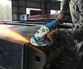 Особа која носи заштитну опрему бруси ивицу челичне греде угаоном брусилицом.