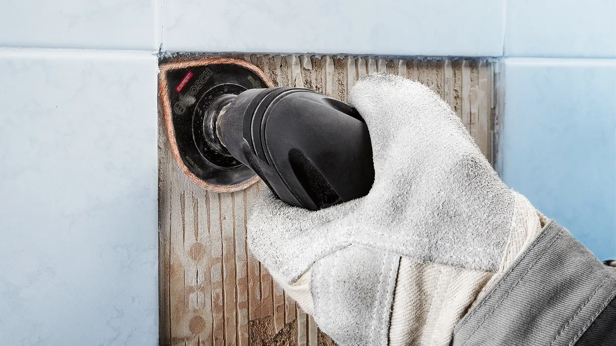 Особа која носи заштитну опрему уклања фугу са плочица помоћу електричног алата.