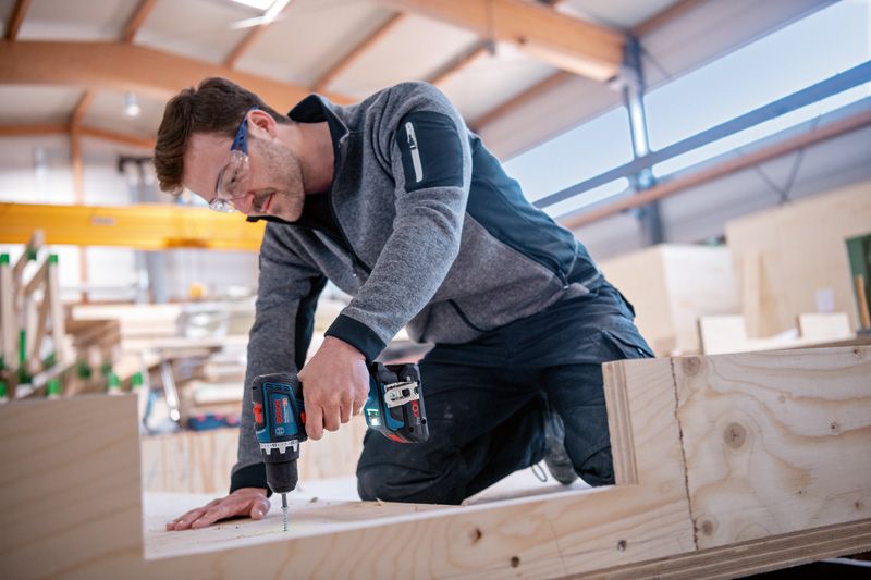 Особа која носи заштитну опрему користи акумулаторску бушилицу да уврне шраф у дрво.