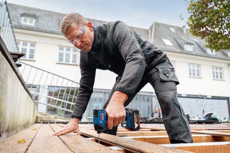 Особа која носи заштитну опрему користи акумулаторску бушилицу за причвршћивање дасака за терасу.